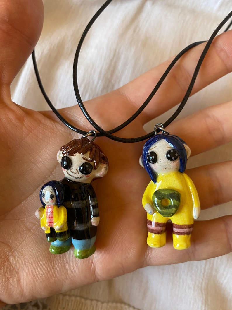 May include: Two handmade pendant necklaces featuring cartoon character figures. One figure has brown hair and a black and gray striped shirt, holding a smaller figure. The other figure has blue hair and a yellow outfit. Both pendants are on black cords.