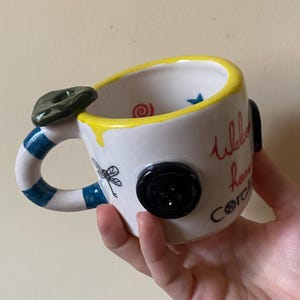 May include: A white ceramic mug with a yellow rim, a blue and white striped handle, and a dark green button accent. The mug features black dragonfly illustrations, a red spiral, a blue star, and the text "Welcome home Coraline" in red and black.