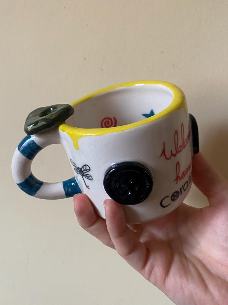 May include: A white ceramic mug with a yellow rim, a blue and white striped handle, and a dark green button accent. The mug features black dragonfly illustrations, a red spiral, a blue star, and the text "Welcome home Coraline" in red and black.
