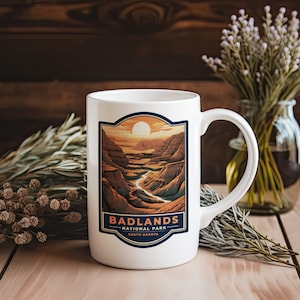 May include: White ceramic mug with a colorful illustration of Badlands National Park in South Dakota. The mug has a handle and says "Badlands National Park, South Dakota" on the front.