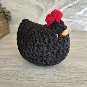 May include: A handmade, black crochet chicken with a red comb and an orange beak. The chicken has a round body and a small, black bead for an eye. This soft sculpture is a charming addition to any home decor.