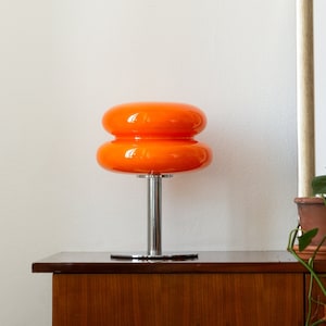 Orange Mid-Century Tischlampe mit Edelstahlfuß und Glasoberfläche auf Kommode neben Pflanze und Bild