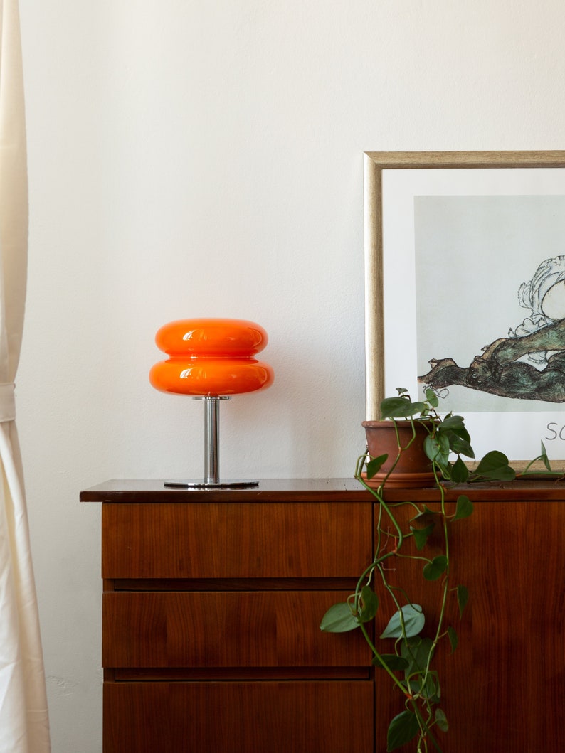 Orange Mid-Century Tischlampe mit Edelstahlfuß und Glasoberfläche auf Kommode neben Pflanze und Bild