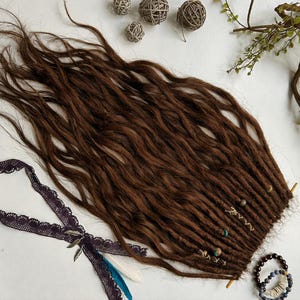 May include: Dark brown dreadlocks with wavy texture, adorned with wooden beads and small decorative elements. A dark purple lace ribbon with feathers and a beaded bracelet are also present. The dreadlocks are arranged on a white surface.