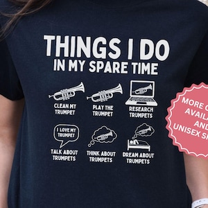 Peut inclure: T-shirt noir avec texte et graphiques blancs. Le texte dit "THINGS I DO IN MY SPARE TIME" avec des illustrations de trompettes et d'activités connexes. Un autocollant rose indique "MORE COLORS AVAILABLE AND UNISEX SIZING".