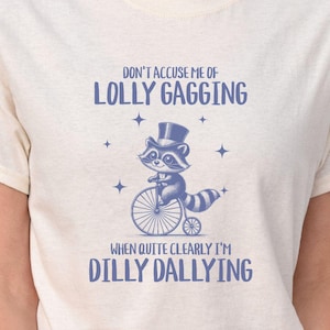 May include: A white t-shirt with a blue raccoon wearing a top hat and riding a bicycle. The text on the shirt reads "Don't accuse me of lolly gagging when quite clearly I'm dilly dallying."