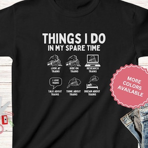 May include: Black t-shirt with white text that reads "Things I do in my spare time" and a graphic of six train-related activities. The graphic includes the text "Look at trains", "Ride on trains", "Research trains", "I love trains", "Talk about trains", "Think about trains", and "Dream about trains". The t-shirt also has a white tag that reads "More colors available".