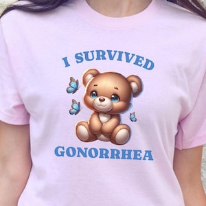May include: Pink t-shirt with a cartoon teddy bear and the text "I survived Gonorrhea" in blue.