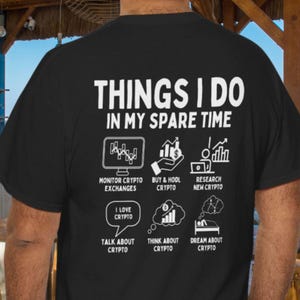 Op de afbeelding: Zwart t-shirt met witte tekst en afbeeldingen. De tekst luidt "THINGS I DO IN MY SPARE TIME" met bijbehorende pictogrammen en zinnen zoals "Monitor Crypto Exchanges" en "Dream About Crypto". Een casual kledingstuk.