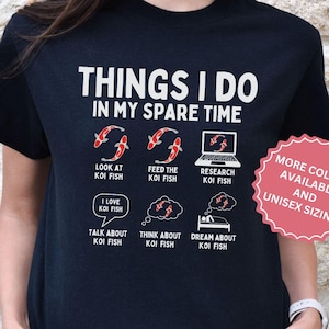 Puede incluir: Camiseta negra con un diseño gráfico blanco que dice "Things I do in my spare time" y presenta ilustraciones de peces koi. Las ilustraciones muestran a una persona mirando peces koi, alimentando peces koi, investigando peces koi, hablando de peces koi, pensando en peces koi y soñando con peces koi. El texto "More colors available and unisex sizing" está en un círculo con un contorno rojo.