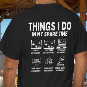 May include: Black t-shirt with white text and graphics. The text reads "THINGS I DO IN MY SPARE TIME" with illustrations of aquariums and related activities. The shirt's design includes phrases like "Look at Aquariums" and "Dream about Aquariums."