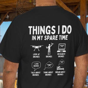 Puede incluir: Camiseta negra con texto blanco y gráficos relacionados con drones. El texto dice "THINGS I DO IN MY SPARE TIME" con ilustraciones de drones y frases como "Look at Drones" y "Dream about Drones".