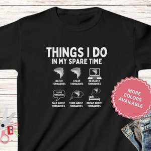 May include: Black t-shirt with a white graphic that says "Things I do in my spare time" and lists activities related to tornadoes. The graphic includes illustrations of tornadoes, a person chasing a tornado, and a laptop. The text "More Colors Available" is in a pink circle on the right side of the image.