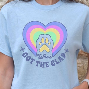 May include: Light blue t-shirt with a rainbow heart and a paw print in the center. The text "Who's Got The Clap" is written below the heart.