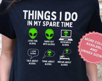 May include: Black t-shirt with a white graphic that says "Things I do in my spare time" and features a series of green alien illustrations. The illustrations depict aliens looking for aliens, hanging out with aliens, researching aliens, talking about aliens, thinking about aliens, and dreaming about aliens. The text "More colors available and unisex sizing" is in a pink circle in the upper right corner.