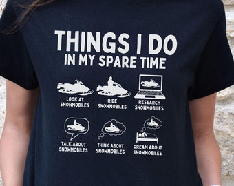 Pode incluir: Uma camiseta preta com texto branco que diz "Things I do in my spare time". O texto é seguido por uma lista de atividades relacionadas a motos de neve, incluindo "look at snowmobiles", "ride snowmobiles", "research snowmobiles", "talk about snowmobiles", "think about snowmobiles" e "dream about snowmobiles".