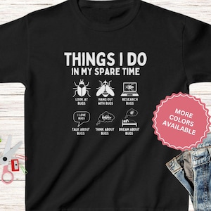 May include: Black t-shirt with white text that reads "Things I do in my spare time". The text is surrounded by illustrations of bugs and activities related to bugs. The text "More Colors Available" is in a pink circle.