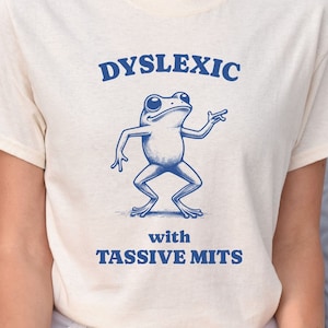 May include: A white t-shirt with a blue frog illustration and the text "Dyslexic with Tassivve Mits".