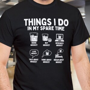 Könnte beinhalten: Ein schwarzes T-Shirt mit weißem Text, der "Things I do in my spare time" lautet. Der Text ist umgeben von Illustrationen von Gläsern Whisky, einer Flasche Whisky, einem Laptop, einem Herzen mit den Worten "I love whiskey", einer Gedankenblase mit einem Glas Whisky und einem Bett mit einer schlafenden Person.