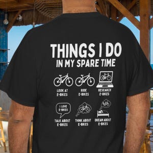 May include: A black t-shirt featuring white text and graphics on the back. The text reads THINGS I DO IN MY SPARE TIME, followed by six icons with labels: LOOK AT E-BIKES, RIDE E-BIKES, RESEARCH E-BIKES, TALK ABOUT E-BIKES, THINK ABOUT E-BIKES, and DREAM ABOUT E-BIKES.