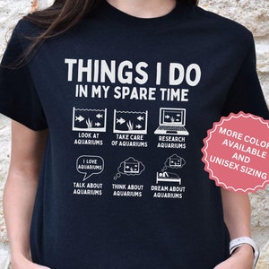 Op de afbeelding: Een zwart T-shirt met een witte afbeelding waarop staat "Things I do in my spare time" en activiteiten worden opgesomd die te maken hebben met aquaria. De afbeelding bevat illustraties van aquaria, een computer en gedachtebellen. De tekst "More colors available and unisex sizing" staat in een roze cirkel aan de rechterkant van het shirt.