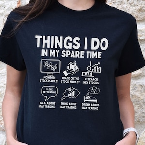 May include: A black t-shirt with white text that reads "Things I do in my spare time". The text is surrounded by icons related to stock market trading, including a computer monitor, a hand holding a dollar bill, a graph, a speech bubble with the text "I love day trading", a coin with a dollar sign, a bed, and a speech bubble with the text "Talk about day trading".
