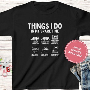 May include: Black t-shirt with a white graphic that says "Things I do in my spare time" and features a list of activities related to armadillos. The graphic includes illustrations of armadillos, a laptop, and a bed. The text "More Colors Available" is in a pink circle.