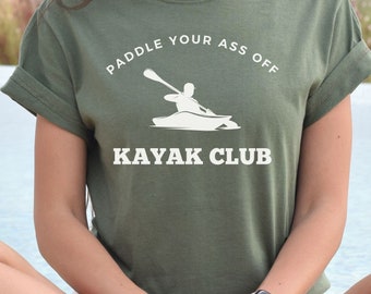 Puede incluir: Una mujer con una camiseta verde con el texto "Paddle Your Ass Off Kayak Club" y una silueta blanca de una persona haciendo kayak.