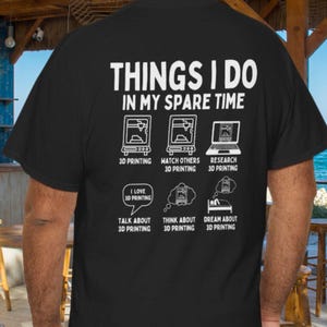 以下が含まれることがあります： 3Dプリントに関する白文字とグラフィックが特徴の黒いTシャツ。「THINGS I DO IN MY SPARE TIME」のテキストと3Dプリンターや関連アクティビティのイラスト。カジュアルウェアアイテムです。