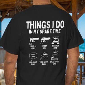May include: Black t-shirt with white text and graphics. The text reads "THINGS I DO IN MY SPARE TIME" with illustrations of guns and a laptop. The shirt also includes phrases like "Look at Guns" and "Dream about Guns".
