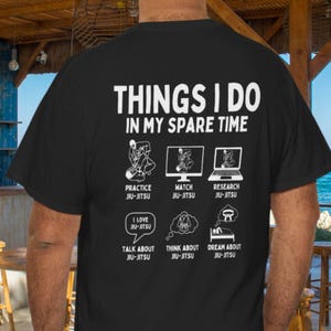 May include: Black t-shirt with white text and graphics. The text reads "THINGS I DO IN MY SPARE TIME" with illustrations of Jiu-Jitsu activities: practicing, watching, researching, loving, talking, thinking, and dreaming about it.