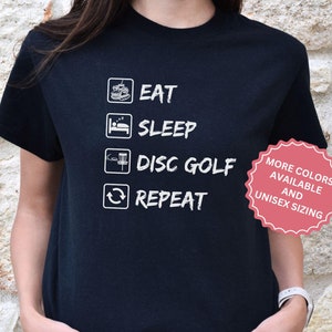 May include: Black t-shirt with white text that reads "Eat, Sleep, Disc Golf, Repeat". The shirt also has a small graphic of a disc golf basket and a disc. A red circle with white text says "More Colors Available and Unisex Sizing".