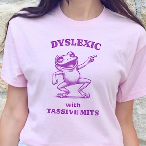 May include: Pink t-shirt with a purple frog graphic and the text "Dyslexic with Tassiive Mits".