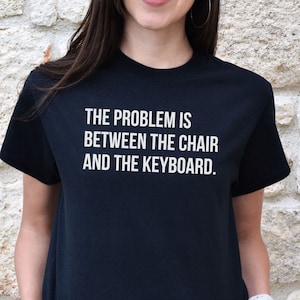 May include: Black t-shirt with white text that reads "The problem is between the chair and the keyboard."