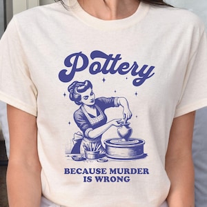 May include: A white t-shirt with a blue graphic of a woman making pottery on a wheel. The text "Pottery" is in a blue retro font above the image. The text "Because murder is wrong" is below the image.
