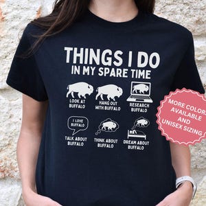 May include: Black t-shirt with white text and buffalo illustrations. The shirt reads "THINGS I DO IN MY SPARE TIME" with various buffalo-related activities. A pink sticker indicates more colors and unisex sizing are available.