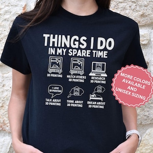 以下が含まれることがあります： 3Dプリントに関する白文字とグラフィックが特徴の黒いTシャツ。「THINGS I DO IN MY SPARE TIME」の文字と3Dプリンターや関連アクティビティのイラストが描かれています。ピンクのステッカーは、より多くの色とユニセックスサイズを示しています。