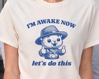 May include: A white t-shirt with a blue graphic of a cartoon bear wearing a hat and overalls. The text "I'm awake now let's do this" is printed in blue on the shirt.
