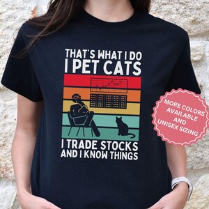 Może przedstawiać: Czarna koszulka z napisem "THAT'S WHAT I DO I PET CATS I TRADE STOCKS AND I KNOW THINGS". Grafika przedstawia osobę handlującą akcjami, kota i schemat kolorów retro. Koszulka ma również różową metkę z napisem "WIĘCEJ KOLORÓW DOSTĘPNYCH I ROZMIARY UNISEX."