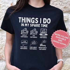 May include: Black t-shirt with white text that reads "Things I do in my spare time". The text is surrounded by illustrations of ham radios, a computer, and thought bubbles with text that reads "I love ham radios", "Talk about ham radios", "Think about ham radios", and "Dream about ham radios". A pink circle with white text that reads "More colors available and unisex sizing" is in the upper right corner.