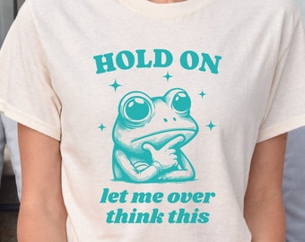 May include: A white t-shirt with a teal green frog graphic and the text "HOLD ON let me over think this".