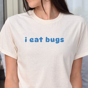 May include: A cream-coloured t-shirt with the blue text "i eat bugs". The short-sleeved shirt is made of a soft material. The shirt is a casual wear item.