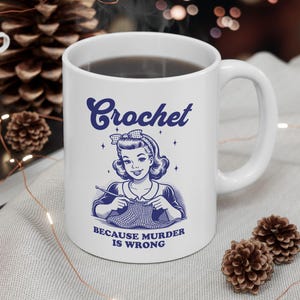 May include: White ceramic mug with a blue illustration of a woman crocheting. The mug has the text "Crochet because murder is wrong" printed on it.