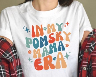 Könnte beinhalten: Weißes T-Shirt mit einer bunten Grafik im Retro-Stil, auf dem "In My Pomsky Mama Era" mit Pfotenabdrücken und Sternen steht.