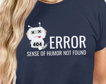 Puede incluir: Una camiseta azul marino con un gráfico de robot blanco y el texto "ERROR SENSE OF HUMOR NOT FOUND". El robot tiene una cara blanca con dos X negras para los ojos y una lengua rosa que sobresale. El robot tiene el número 404 en su pecho.