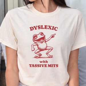 May include: A white t-shirt with a red graphic of a frog dancing and the text "Dyslexic with Tassivve Mits".
