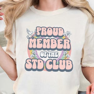 May include: Cream-colored t-shirt with the text "PROUD MEMBER OF THE STD CLUB" in a retro font. The design includes a rainbow, flowers, and a dove. The shirt is a casual, unisex style.