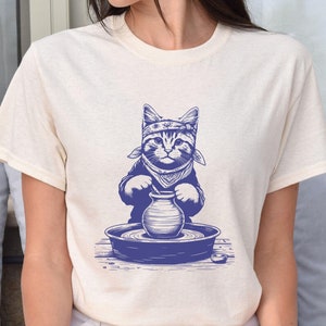 May include: A white t-shirt with a blue illustration of a cat wearing a bandana and making pottery on a pottery wheel.