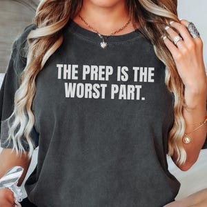 May include: Dark gray t-shirt with the text "THE PREP IS THE WORST PART" in white block letters. The shirt has short sleeves and a relaxed fit. The model is wearing the shirt.