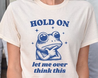 May include: A white t-shirt with a blue frog graphic and the text "HOLD ON let me over think this".